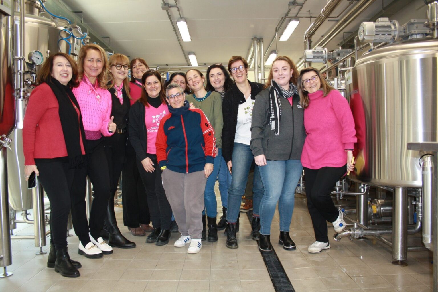 Foto di gruppo delle donne dell’Associazione Le Donne della Birra all’interno di un birrificio, tra fermentatori e impianti di produzione.