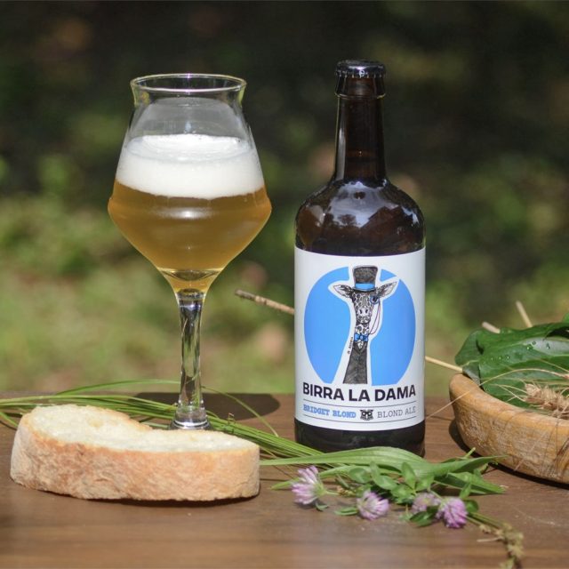 Bottiglia di birra "Birra La Dama" con bicchiere pieno, fetta di pane e fiorellini viola.