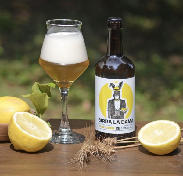 Bottiglia di birra "Birra La Dama" con bicchiere pieno, limoni freschi e spighe di grano.