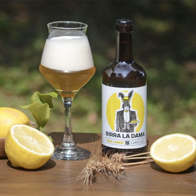 Bottiglia di birra "Birra La Dama" con bicchiere pieno, limoni freschi e spighe di grano.
