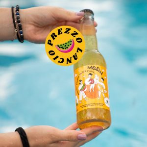 Mani che tengono una bottiglia di bevanda Mesh Orange all'arancia e frutti tropicali davanti a una piscina.